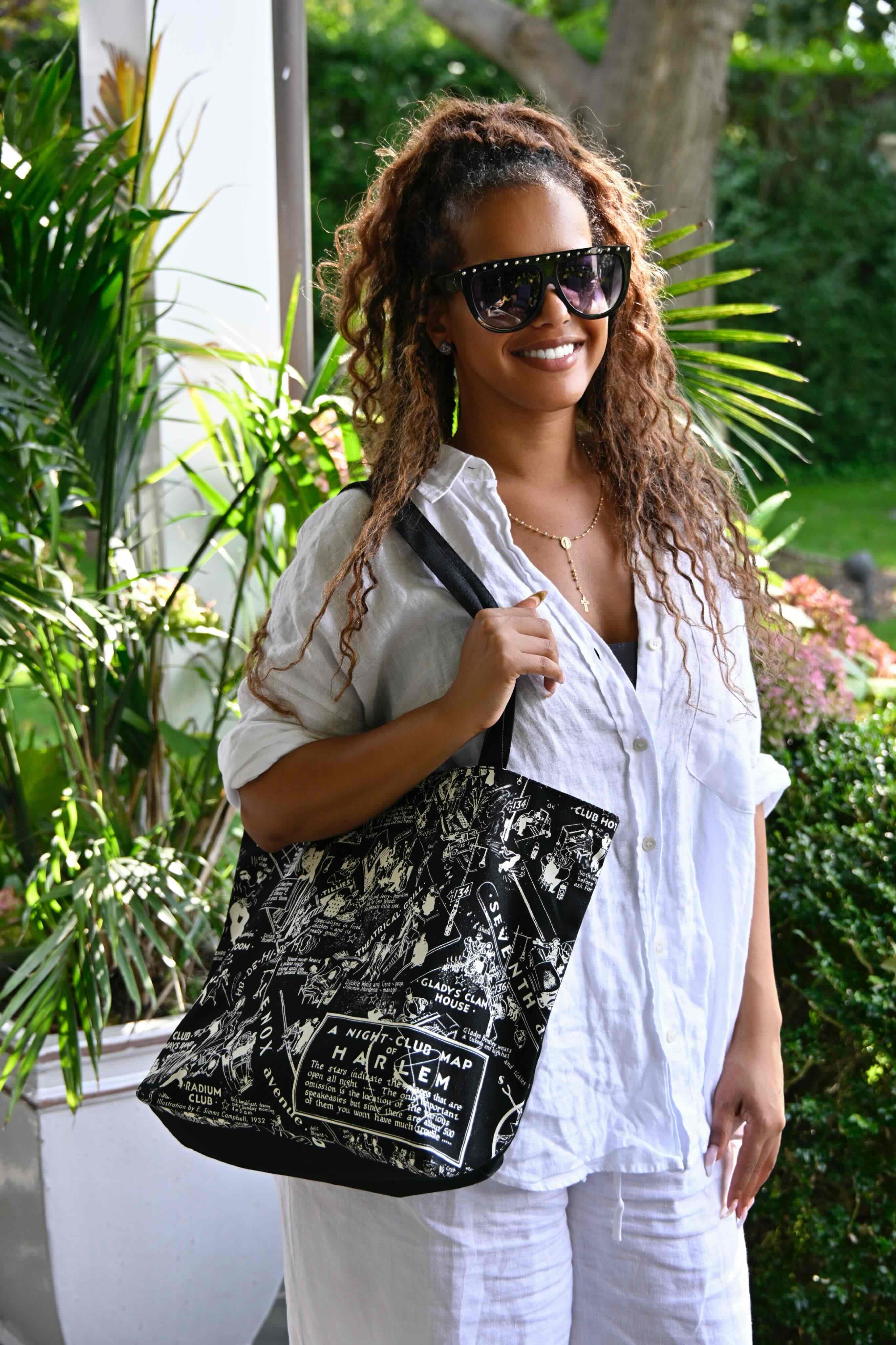 This is an image of a model dressed in white with sunglasses showcasing the black and white Nightclub Map of Harlem tote bag.  