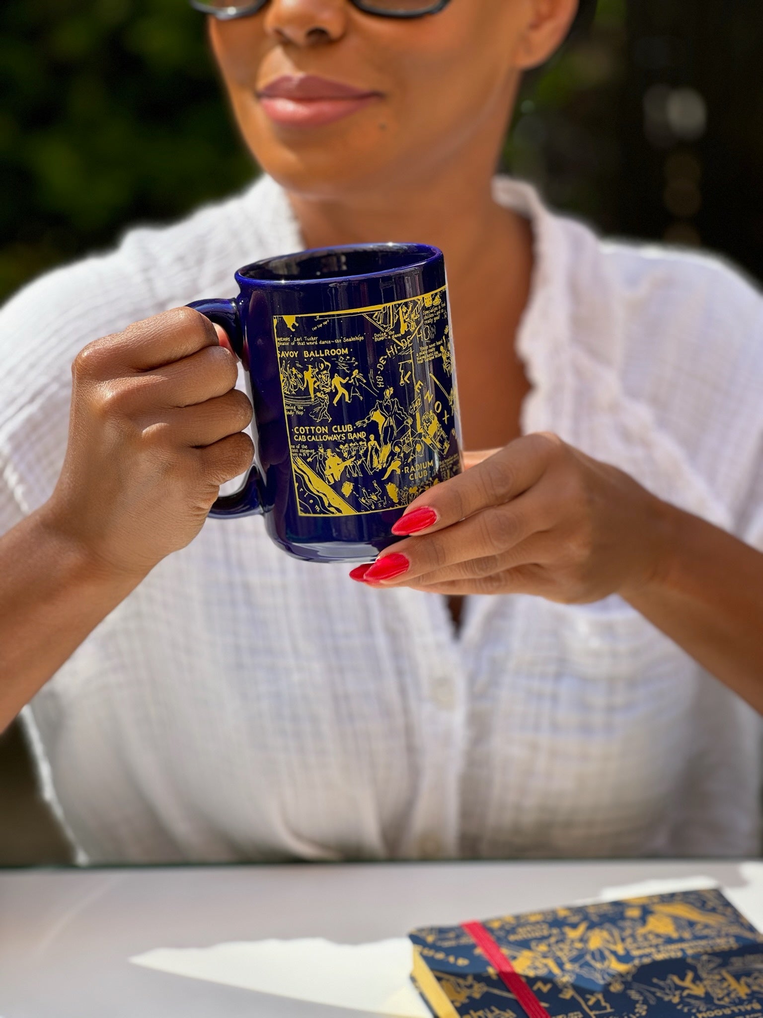 This is a lifestyle image of our blue Nightclub Map of Harlem mug held by a model with red fingernails .  She is sitting in front of the blue nightclub map of Harlem journal