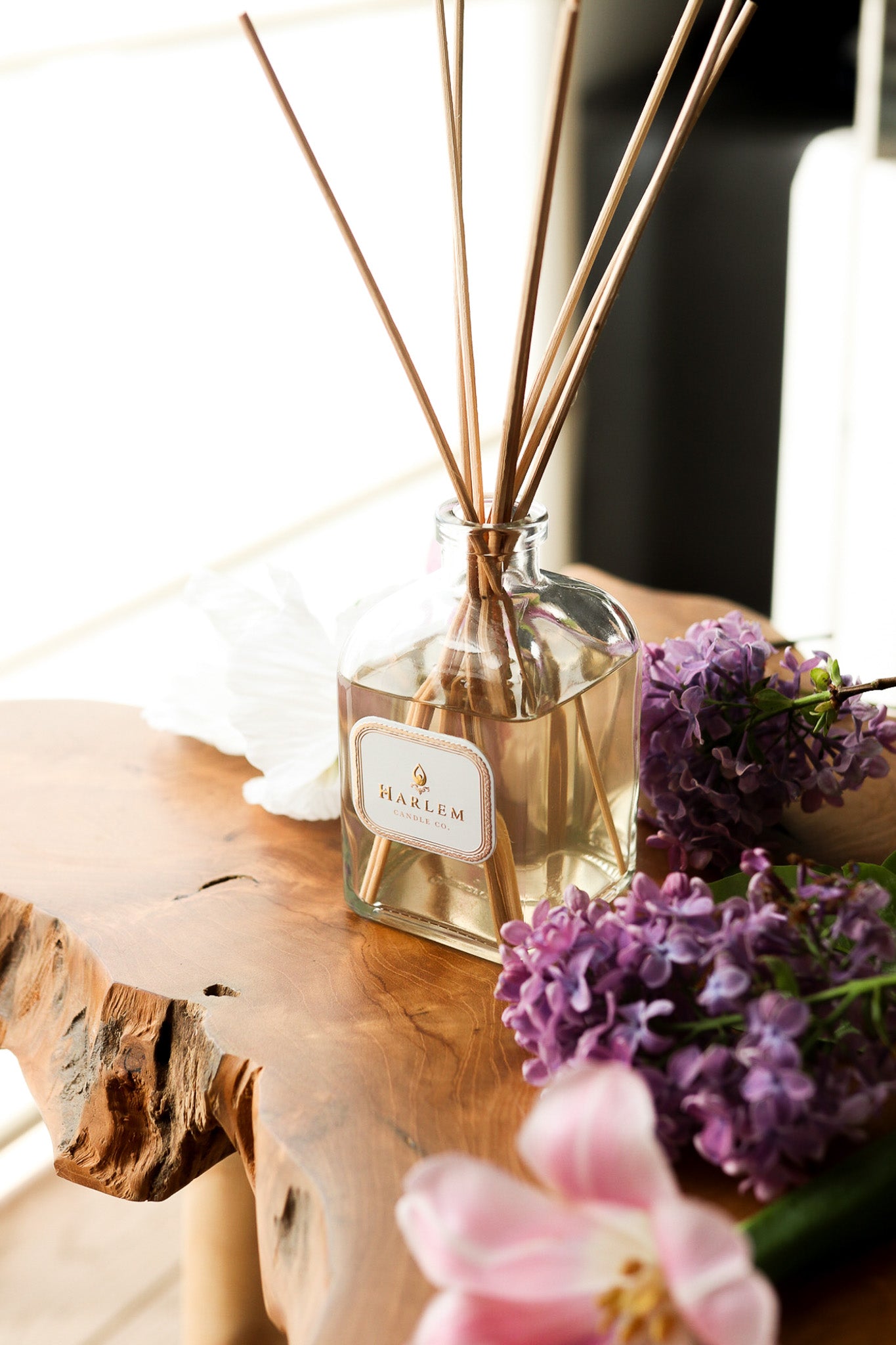 This is a vertical image of the clear glass "Joy" Botanical Diffuser bottle with colorful flowers in the background.