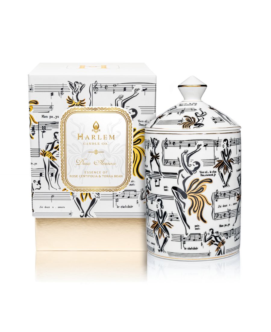 This is an image of our gold and white Deux Amours, ceramic candle with a lid. It features illustrations of Josephine Baker, and other original illustrations.  This candle is featured next to its decorative box.