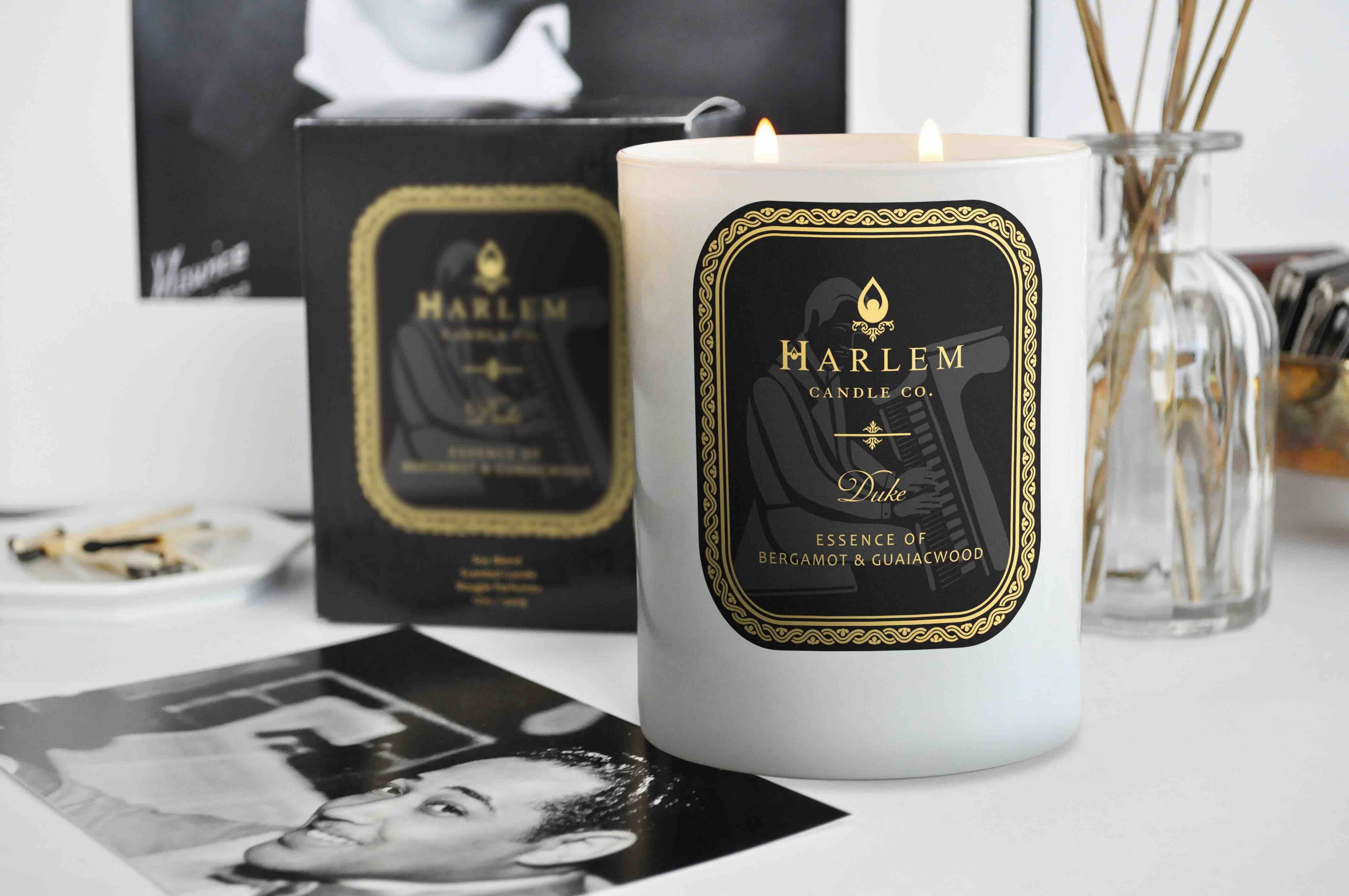 White 11 oz Duke candle with gold and Black label. This candle has 2 wick.  It is a lifestyle picture on a table with pictures of Duke Ellington around. 