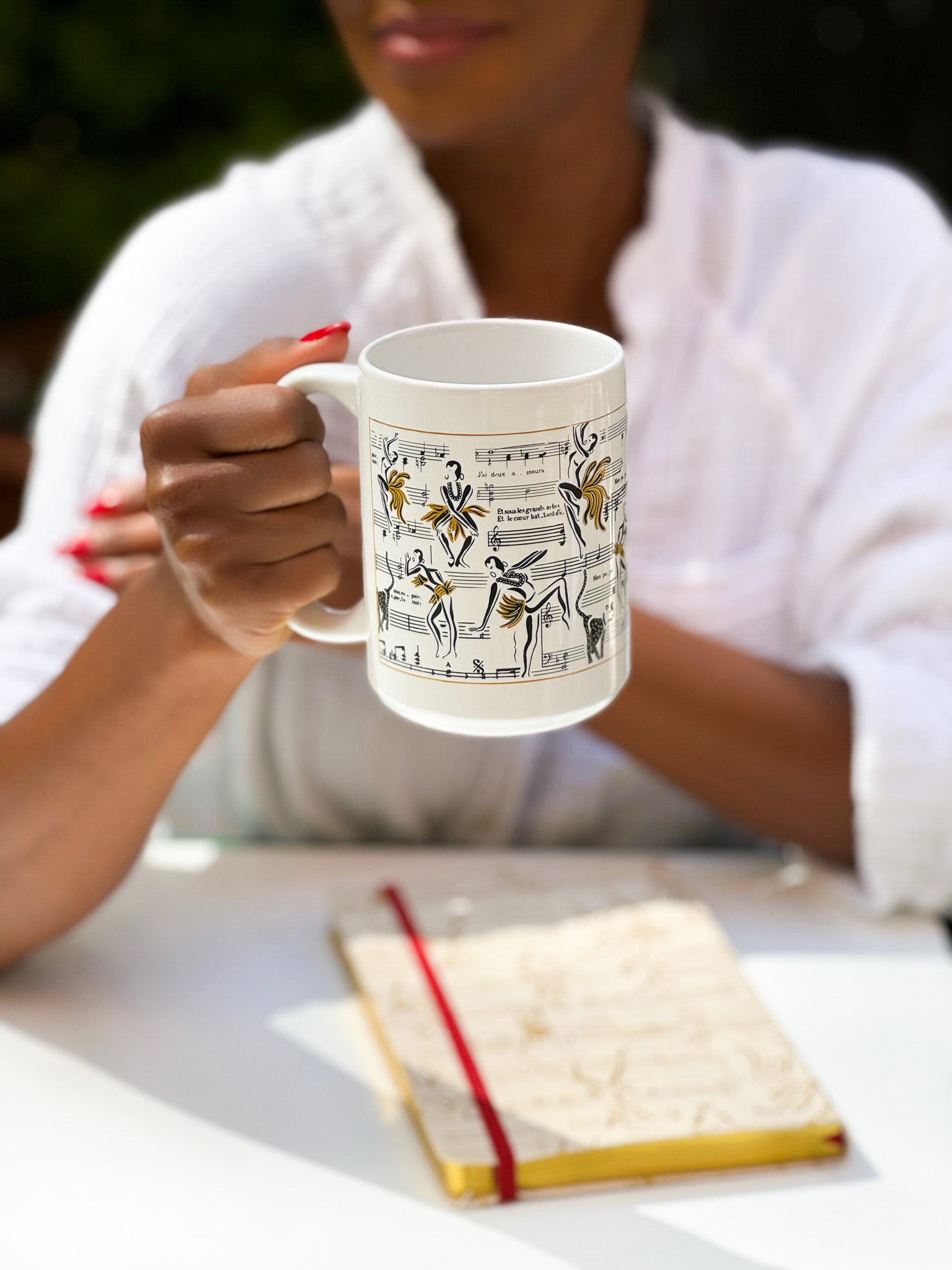 This is an image of the white Josephine mug with illustrations of Josephine Baker dance with her music notes in the background. The Josephine journal is also featured in this image.