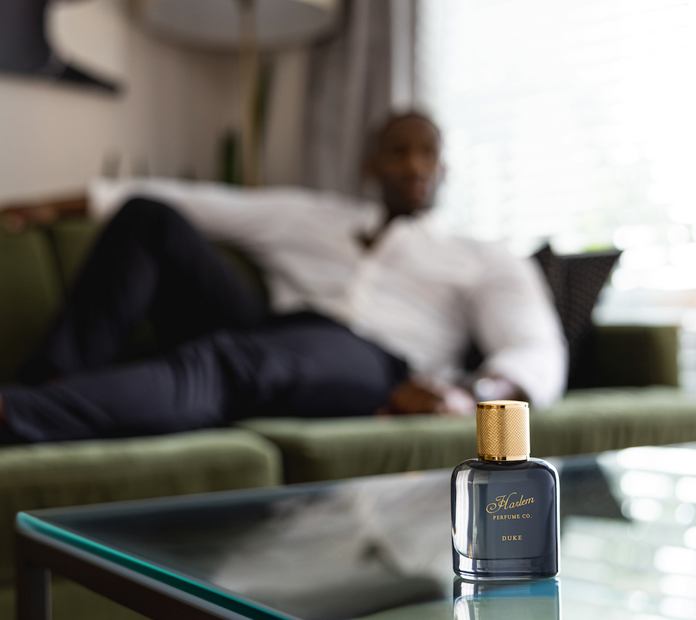 This is an image of our Duke perfume placed on a glass table with a male model, wearing a white shirt and blue pants, leaning casually on a green velvet couch.