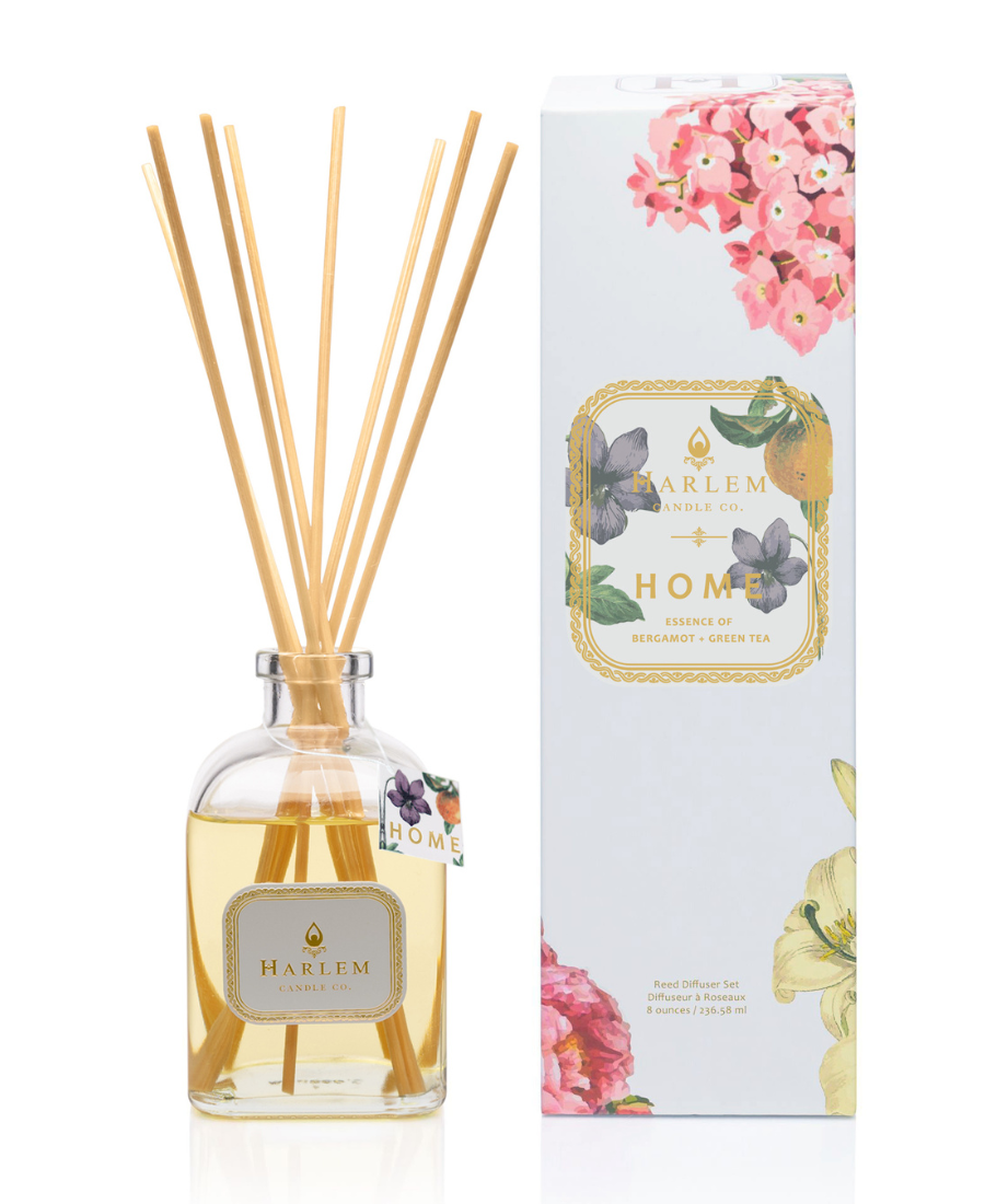 This is an image of our Home reed diffuser in a clear bottle with a white metallic label positioned next to the white floral decorative box.  The diffuser has a hang tag that says Home to indicate that it is different from the other Botanical diffusers.