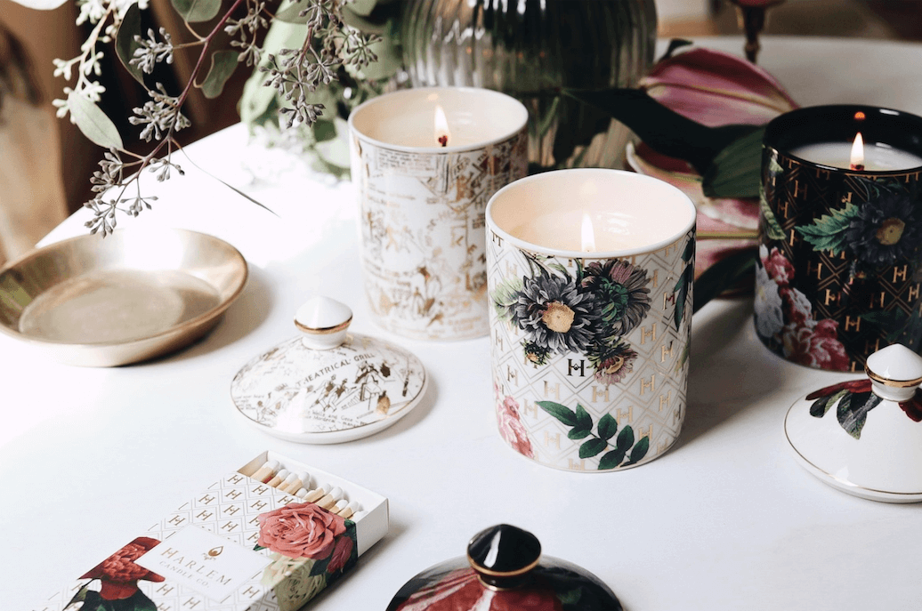 A lifestyle image all three of our lit Ceramic Gold Collection candles: the “Love” by James Baldwin Ceramic Luxury Candle, the “Lady Day” White Floral Ceramic Luxury Candle, and the “Speakeasy” Harlem Map Ceramic Luxury Candle.  Our matches, and a vase with greenery and flowers are on the table as well.