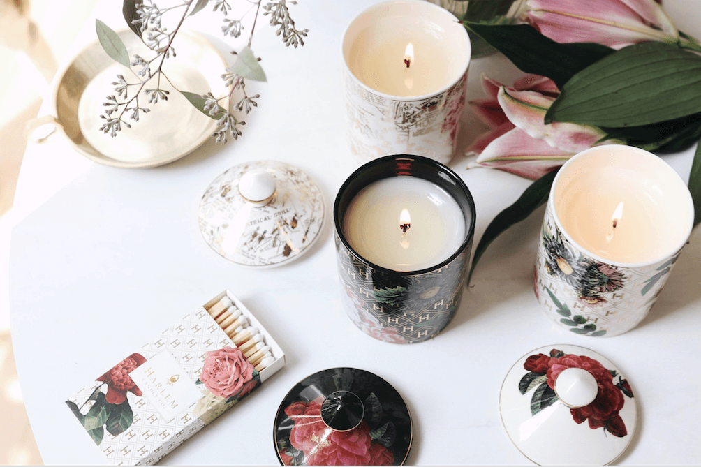 A lifestyle image of all three of our lit Ceramic Gold Collection candles: the “Love” by James Baldwin Ceramic Luxury Candle, the “Lady Day” White Floral Ceramic Luxury Candle, and the “Speakeasy” Harlem Map Ceramic Luxury Candle on a white table with flowers, and the lids of the ceramics sitting nearby.