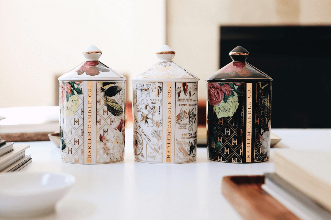 All three of our Ceramic Gold Collection candles: the “Love” by James Baldwin Ceramic Luxury Candle, the “Lady Day” White Floral Ceramic Luxury Candle, and the “Speakeasy” Harlem Map Ceramic Luxury Candle.  A lifestyle image on a white table with books and wooden trays.