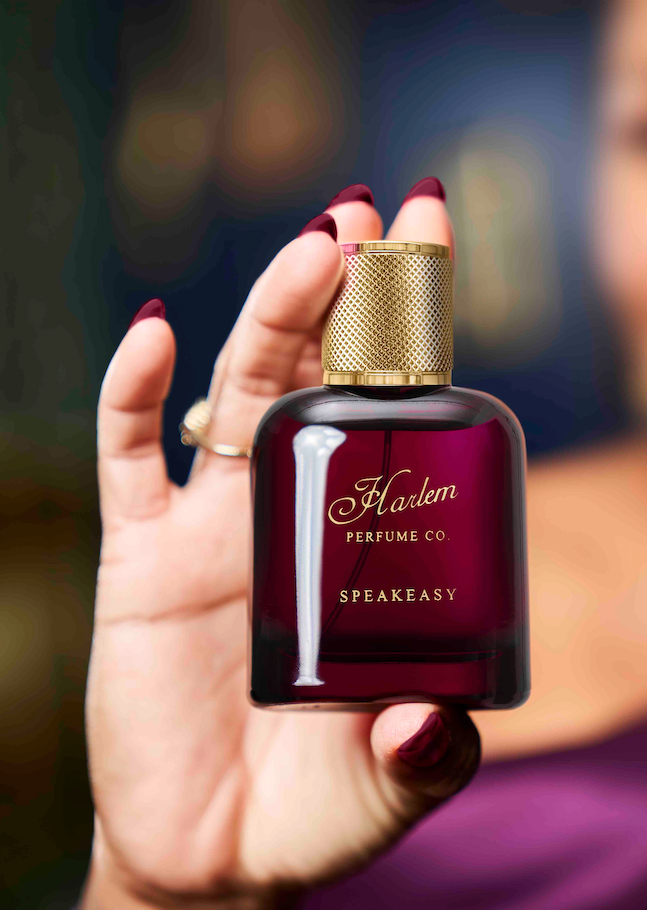 A lifestyle shot of our 50 ml / 1.7 oz Speakeasy eau de parfum held with four fingers in a rich burgundy bottle with a gold cap and the  Harlem Perfume Co logo on the bottle with the word Speakeasy in all caps in a block print font.